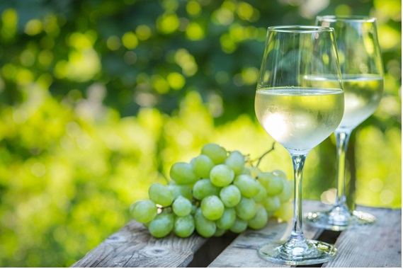 屋外の木製の机の上に白ブドウの房と白ワインのグラスが置かれている画像